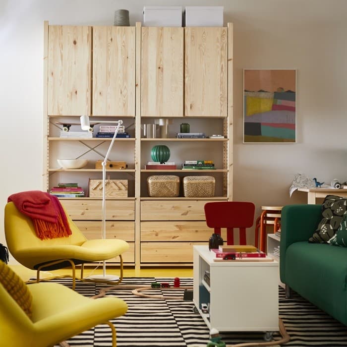 A living room with a natural wood storage unit against the wall, a green sofa, a yellow armchair draped with a red blanket, a striped rug, a white coffee table, and colourful artwork on the wall, creating a warm and inviting atmosphere.