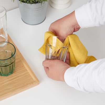 Person wiping glasses with yellow cloth on white table. Two glasses, plant, and wooden board nearby.